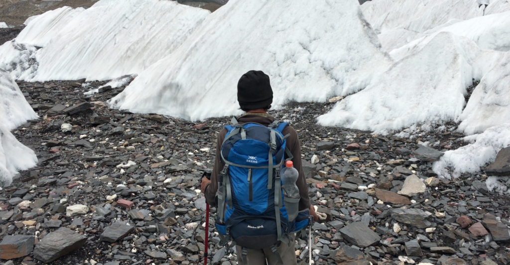 Trekker with a backpack and trekking poles walking over rocky moraine beside ice seracs on the Baltoro Glacier.