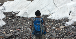 Trekker with a backpack and trekking poles walking over rocky moraine beside ice seracs on the Baltoro Glacier.