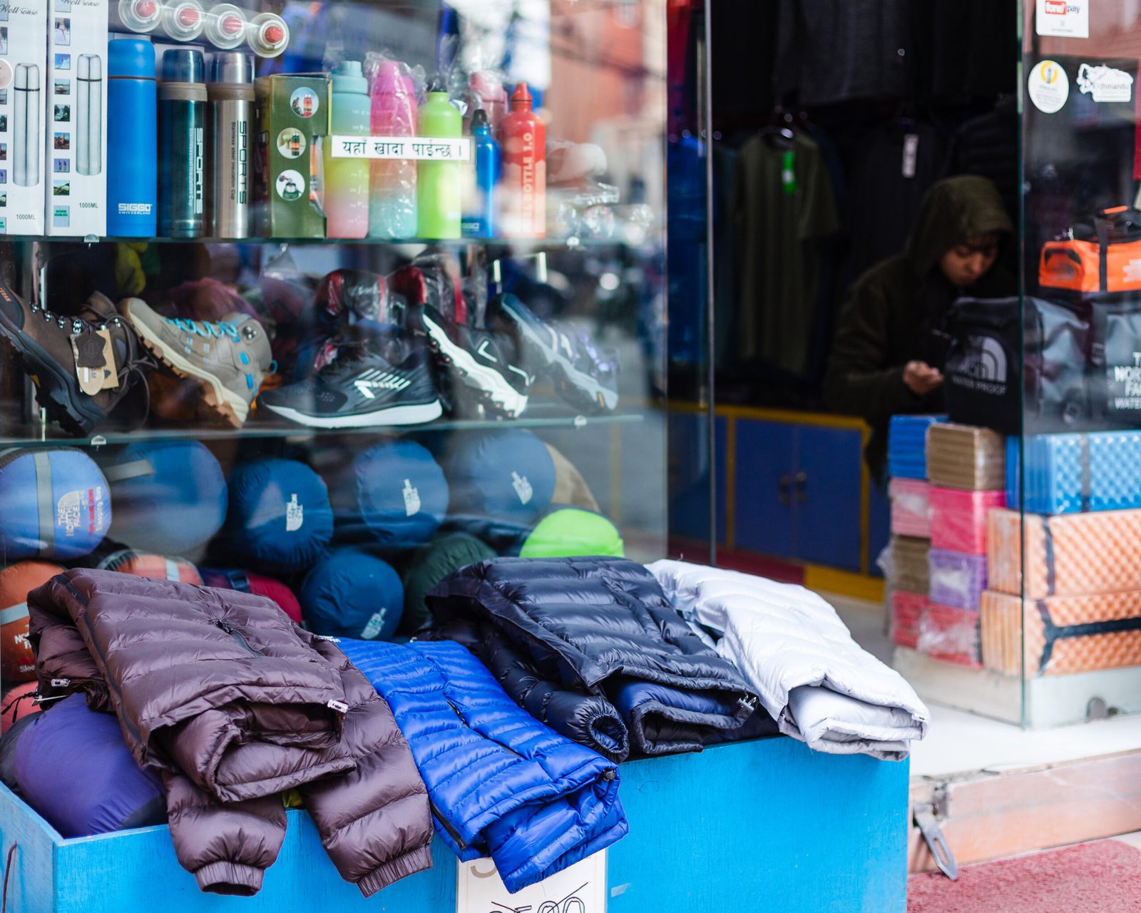 Quilted down jackets laid out outside a trekking shop in Thamel, Kathmandu, with boots and bottles behind.