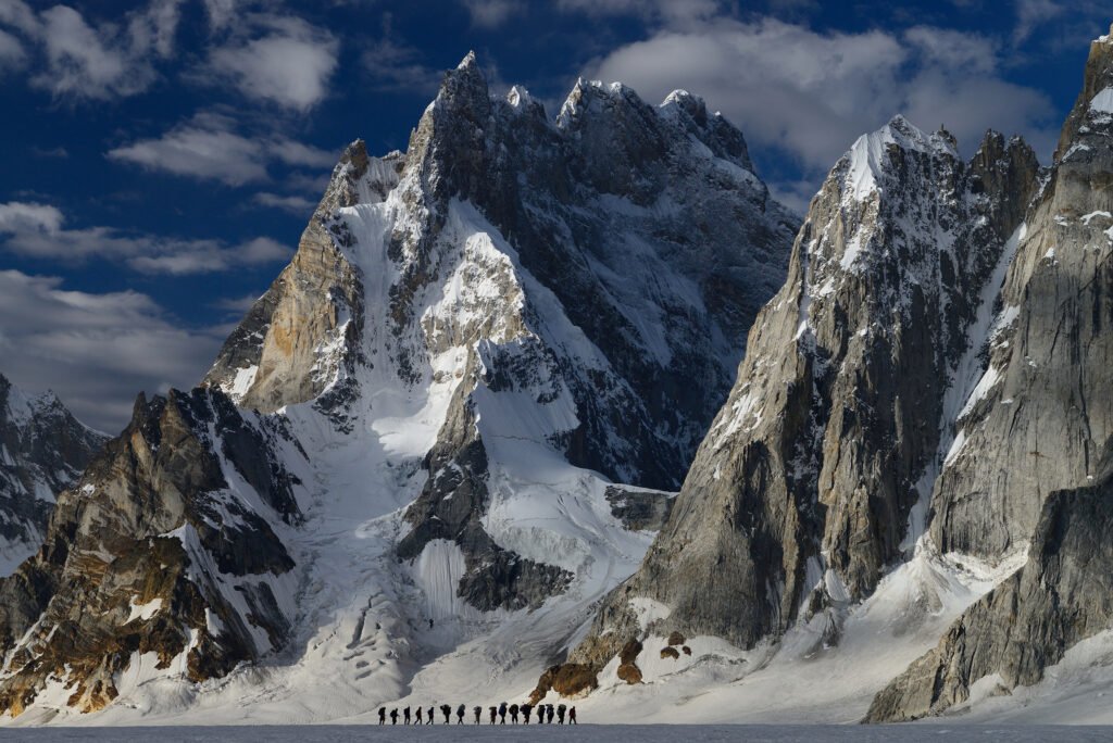 Trekkers and porters crossing a snowfield beneath steep peaks towards Snow Lake on the Biafo Glacier